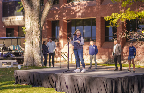 A group of people standing on an outdoor stage in front of a campus building, speaking to an audience gathered on the lawn.
