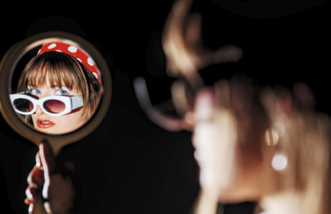 A student model poses with Hollywood style glasses and looks at the camera through the reflection in a handheld mirror