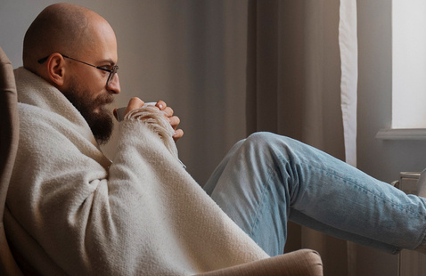 A man in his fifties sits on a sofa, drinking a cup of coffee. He wears a blanket around his arms and shoulders..