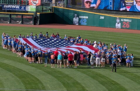 Cancer survivors stand on a baseball field holding a giant U.S. flag. 