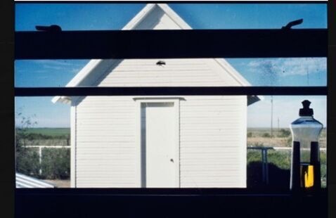 A white shed can be seen through a kitchen window looking out. The window frame can be seen and there is a bottle of detergent on the right side of the counter.
