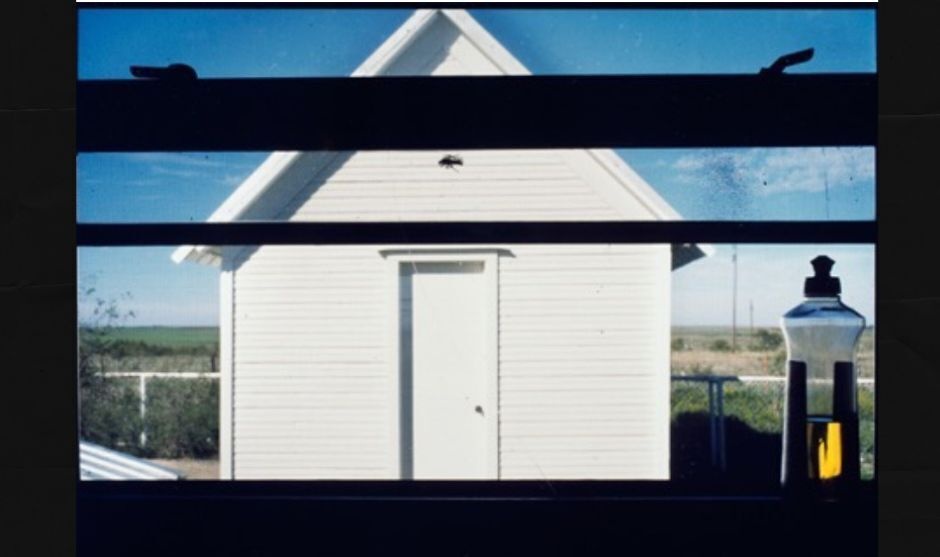 A white shed can be seen through a kitchen window looking out. The window frame can be seen and there is a bottle of detergent on the right side of the counter.