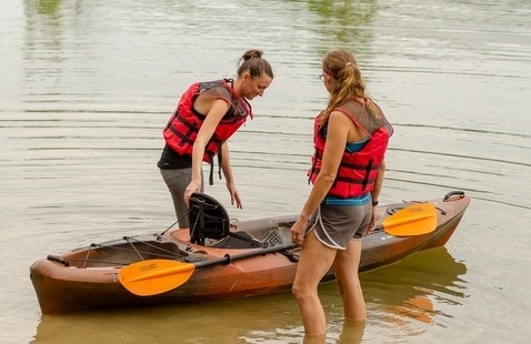 An orange and back kayak floats in shallow water. A paddle is attached to one side of the boat. A woman wearing a life jacket stands on each side of the kayak, with their feet just over ankle-deep in the water.