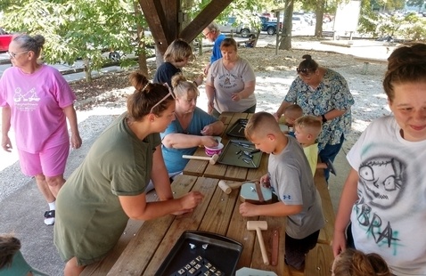 Adults and children work on a craft together at picnic tables underneath a shelter.