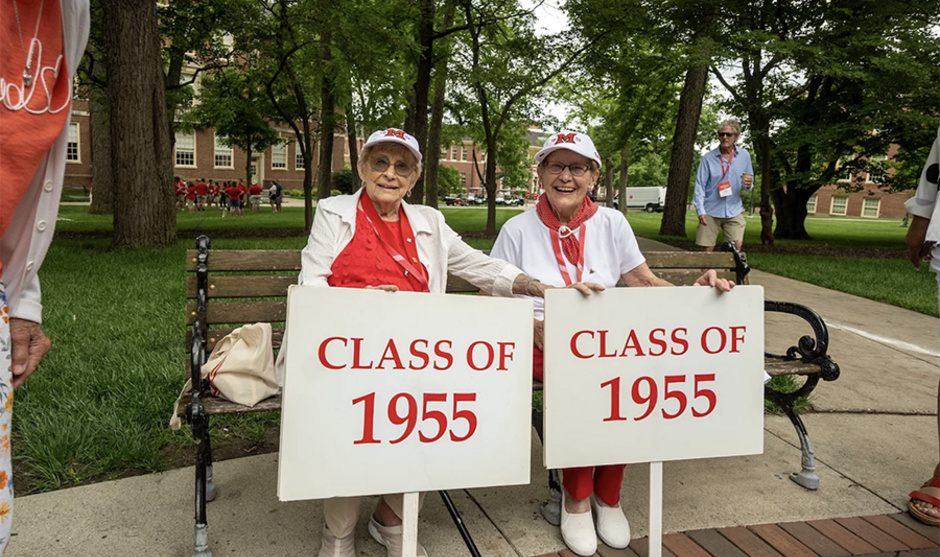 Two women sit on a bench on campus and hold signs that indicate 'class of 1955'