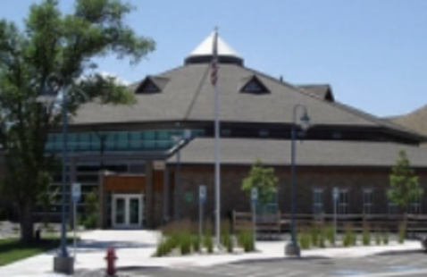 Exterior view of the Spanish Springs Library, brick building with multi-sided roof, large windows, and a central entrance with glass doors. A flagpole stands in front, with trees, landscaping, and a sidewalk along the building.
