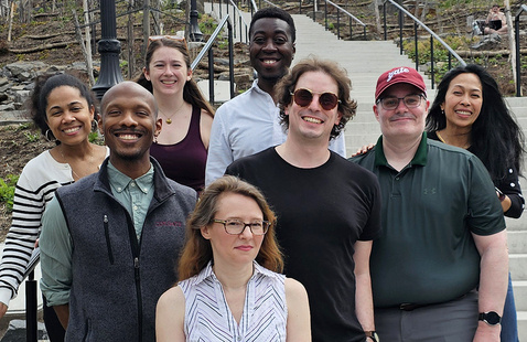 A group of eight diverse individuals are smiling and posing together on and beside an outdoor concrete staircase set into a hillside. The background features a terraced, rocky landscape with young trees and a few distant people relaxing on the slopes.