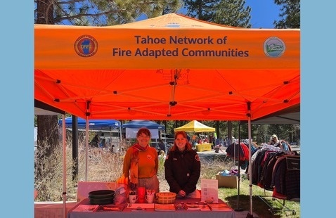 Two women stand behind an information table with a tent above that says Tahoe Network of Fire Adapted Communities with logos for Tahoe Fire and Fuels Team and Tahoe Resource Conservation District.