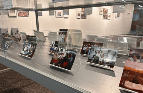 The image shows a glass display case containing several photographs set on clear stands. The case is located in an exhibition space with white walls. Mounted on the back wall are a few framed pictures.