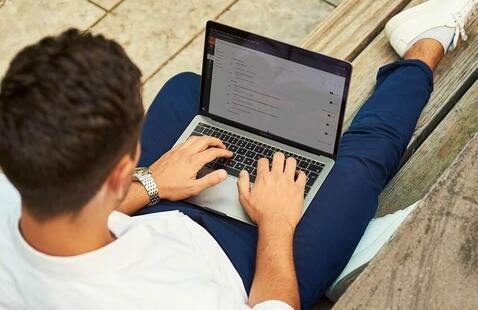 Man lounging on a bench outside with a laptop in his lap.