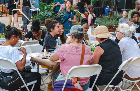 A group of people sit around a table outdoors, talking and crafting at a community event.