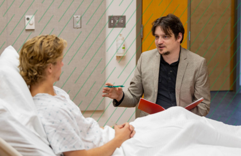 A student sits next to a patient in a hospital setting