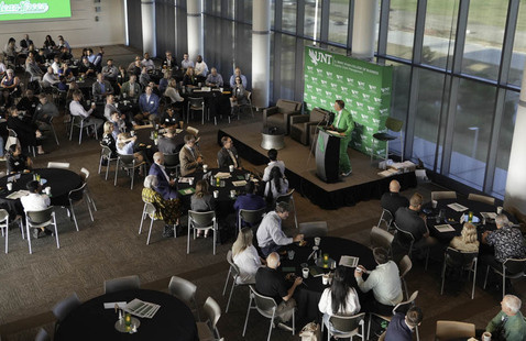 A ballroom, filled with people at tables and a speaker at a stage.