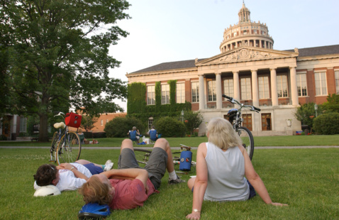 three people lounge on the grass in front of Rush Rhees Library.
