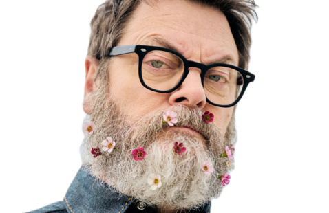 Comedian Nick Offerman a man with short dark hair and glasses looks sternly at the camera wearing a full grey beard dotted with small pink and red wildflowers. 