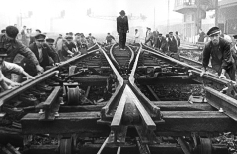 Still from Trains: workers repair converging railroad tracks in a black-and-white archival image, as a lone figure stands above the junction.