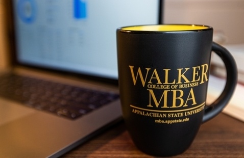 A coffee mug with App State MBA logo sits on a table with a laptop in the background.