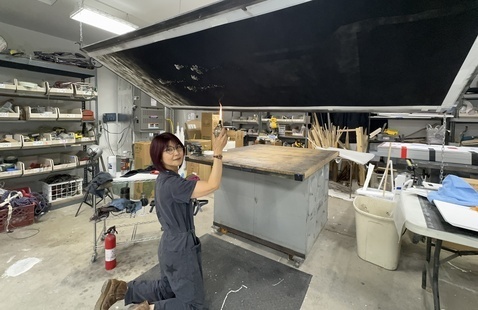 A photograph of Miya Hannan working in her studio. She is wearing a gray jumpsuit, sitting on her knees with her body facing away from the camera. Her face is turned towards the camera and she is holding an object with a flame.