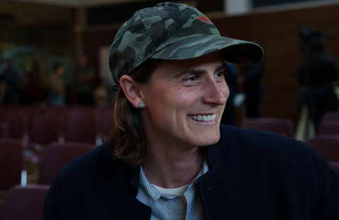 Headshot of Chel Mendell. They are smiling and looking away from the camera while wearing a printed camouflage baseball cap.