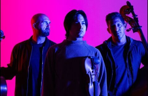 Three string musicians under pink and blue lighting, center holding a violin.