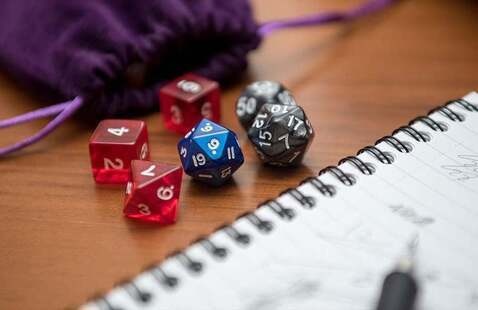 An image of multicolored dice of varying numerical values next to an open notebook with pen and a purple drawstring bag.