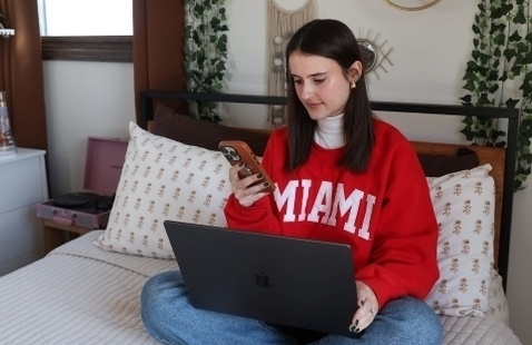 A student in a red Miami sweatshirt with white Miami letters sitting on her bed with a computer in her lap and phone in her hand.