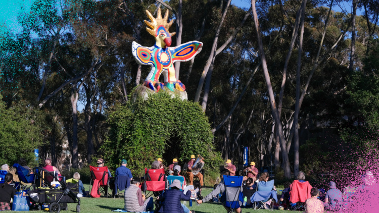 A group of people in lawn chairs sits in front of a large bird sculpture on a grassy lawn. There is a band playing instruments sitting in front of the sculpture. 