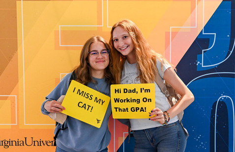 Two students pose with WVU-themed signs in front of a West Virginia University backdrop.