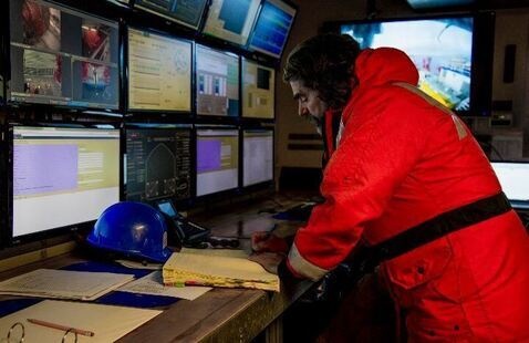 A man in a bright orange jacket stands at a desk in front of many computer screens.