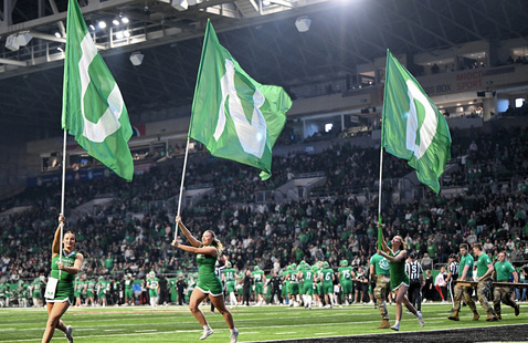 Cheerleaders running across football field carrying three tall flags spelling out UND