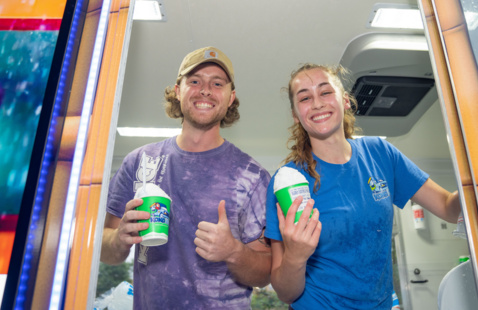Two people inside a snow cone truck serving snow cones and smiling