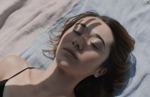 A woman shields her face from the sun with her fingers while lying on a sheet