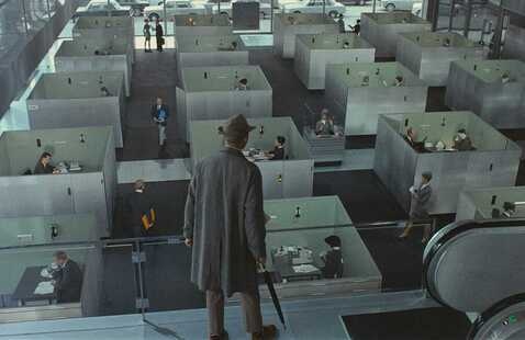 A man in a top hat and long coat looks out over an array of grey cubicle offices