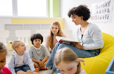 Preschool children gathered around a teacher reading a storybook indoors.