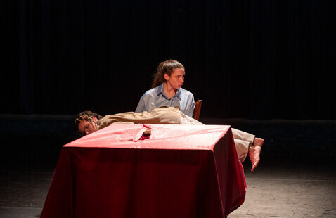 One person is sitting in a chair looking away. Another person is laying on the first person’s lap with their legs extended straight. Both are behind a table covered in a red cloth.