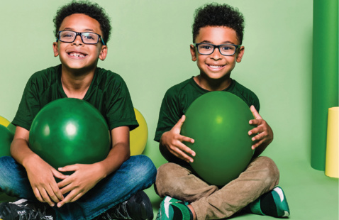 Two children wearing glasses sit on a green studio set holding large green balloons, surrounded by green and yellow balloons.