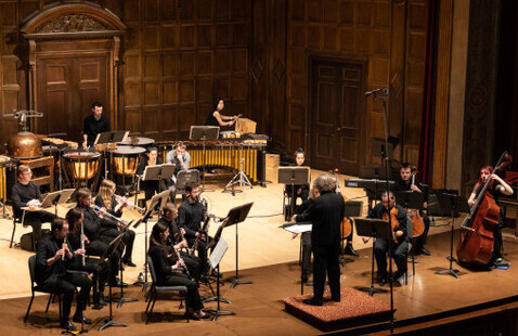 A photo of a music ensemble and conductor performing on a stage with multiple instruments  