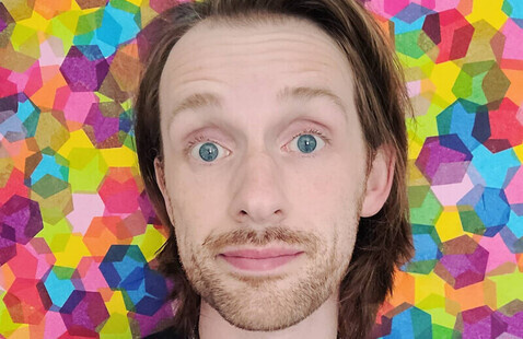 A man looks directly at the camera with eyebrows raised. He’s in front of a colorful, geometric background.