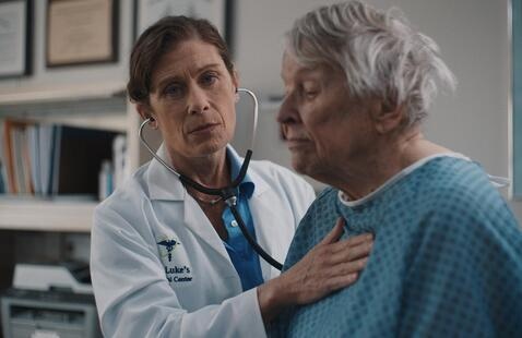 Still from Winter Hymns: a physician examines an elderly patient in a medical office.