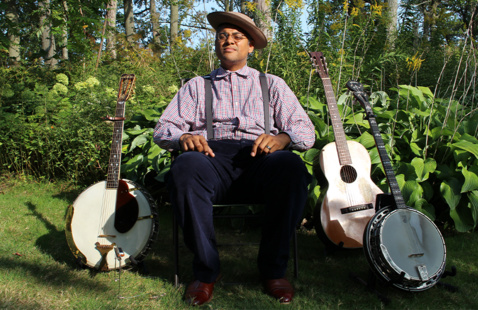 A man sitting on a green lawn with two banjos and a guitar next to him