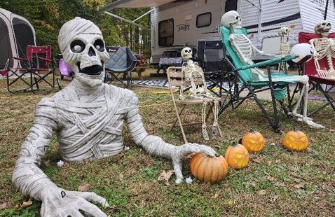 A decorated campsite with a giant mummy emerging from the ground, skeletons sitting in chairs, and glowing jack-o-lanterns.