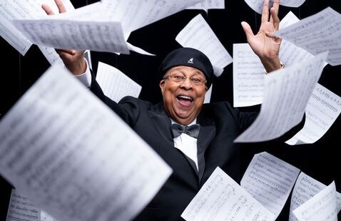 A person wearing a dark suit, bow tie, glasses, and a black hat stands against a black background with both arms raised. Numerous pages of printed sheet music appear to be floating or falling through the air around the person, creating a dynamic composition. The sheets are shown at varying angles and distances from the camera, framing the central figure. Photo credit: Martin Espino Garcia.