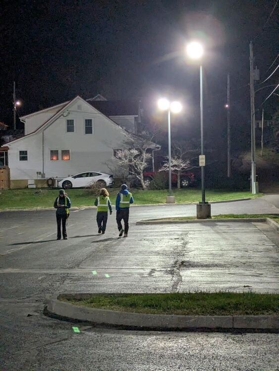 Photo of three students in high visibility vests walking away from the camera, into a large bright street light.
