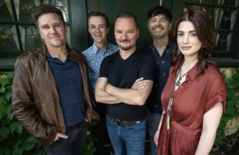 Five band members stand and smile in front of a green bush