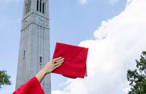 A person holding a graduation hat. The Belltower sits in the background.