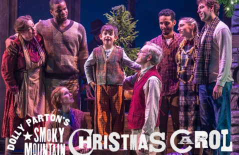 Promotional image for 'Dolly Parton's Smoky Mountain Christmas Carol' showing a diverse cast in rustic period costumes gathered around a young boy on stage, with a small Christmas tree and warm string lights in the background."