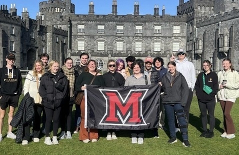 A group of students standing in front of an old stone building holding a black flag with a red "M" on it
