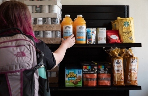 A person with a backpack reaching out to grab a bottle of juice off a food pantry shelf. 