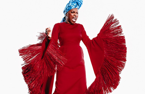 Angelique Kidjo, a woman in a long red dress with extravagantly fringed sleeves and a blue head-wrap, opens her mouth to sing.