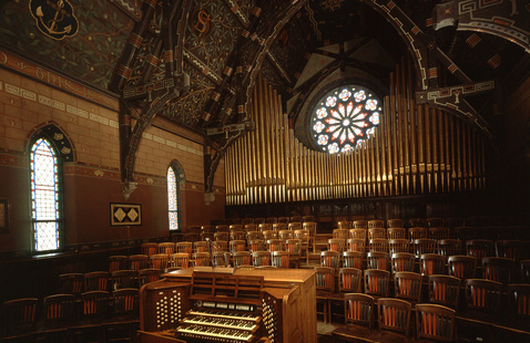 Interior view of a Gothic-style chapel choir loft with rows of wooden chairs, an ornate wooden pipe organ in the foreground, and a large stained-glass rose window above the organ pipes, beneath a richly decorated vaulted ceiling.